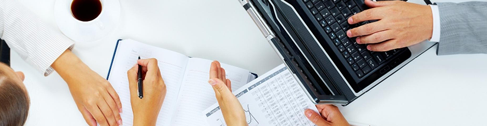 Hands of several people working on ledger books, spreadsheets and a laptop.